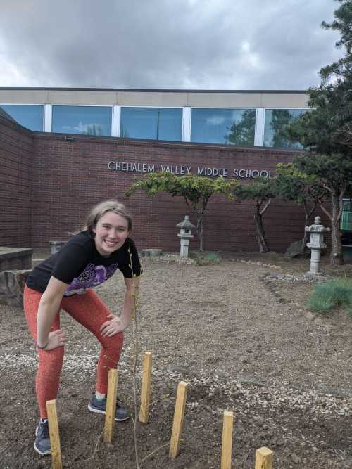 Female student standing behind newly planted tree in front school building